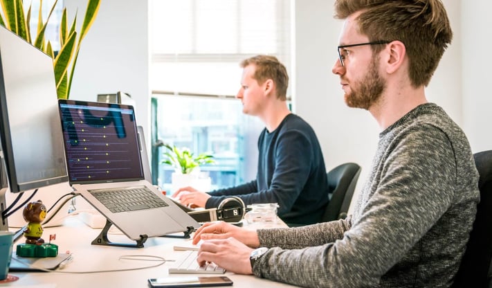 Two guys sitting at a desk with computers and screens